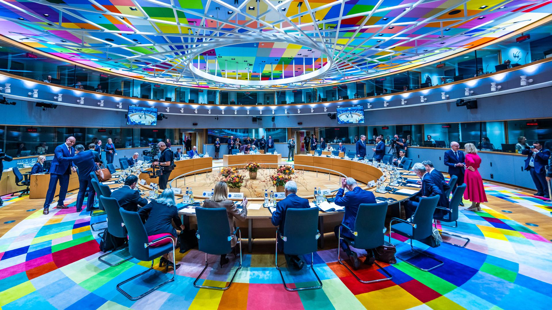 Die Staats- und Regierungschefs der EU-Staaten beraten an einem runden Tisch in Brüssel. Er steht auf einem bunten Teppich, der sich in der Decke spiegelt. 