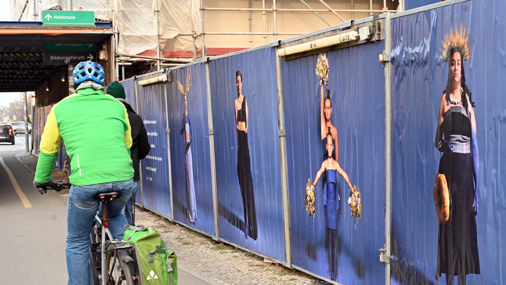 An einem Bauzaun hängen große Poster mit Frauen darauf. Der Hintergrund der Poster ist blau. Ein Fahrradfahrer fährt an dem Bauzaun vorbei.