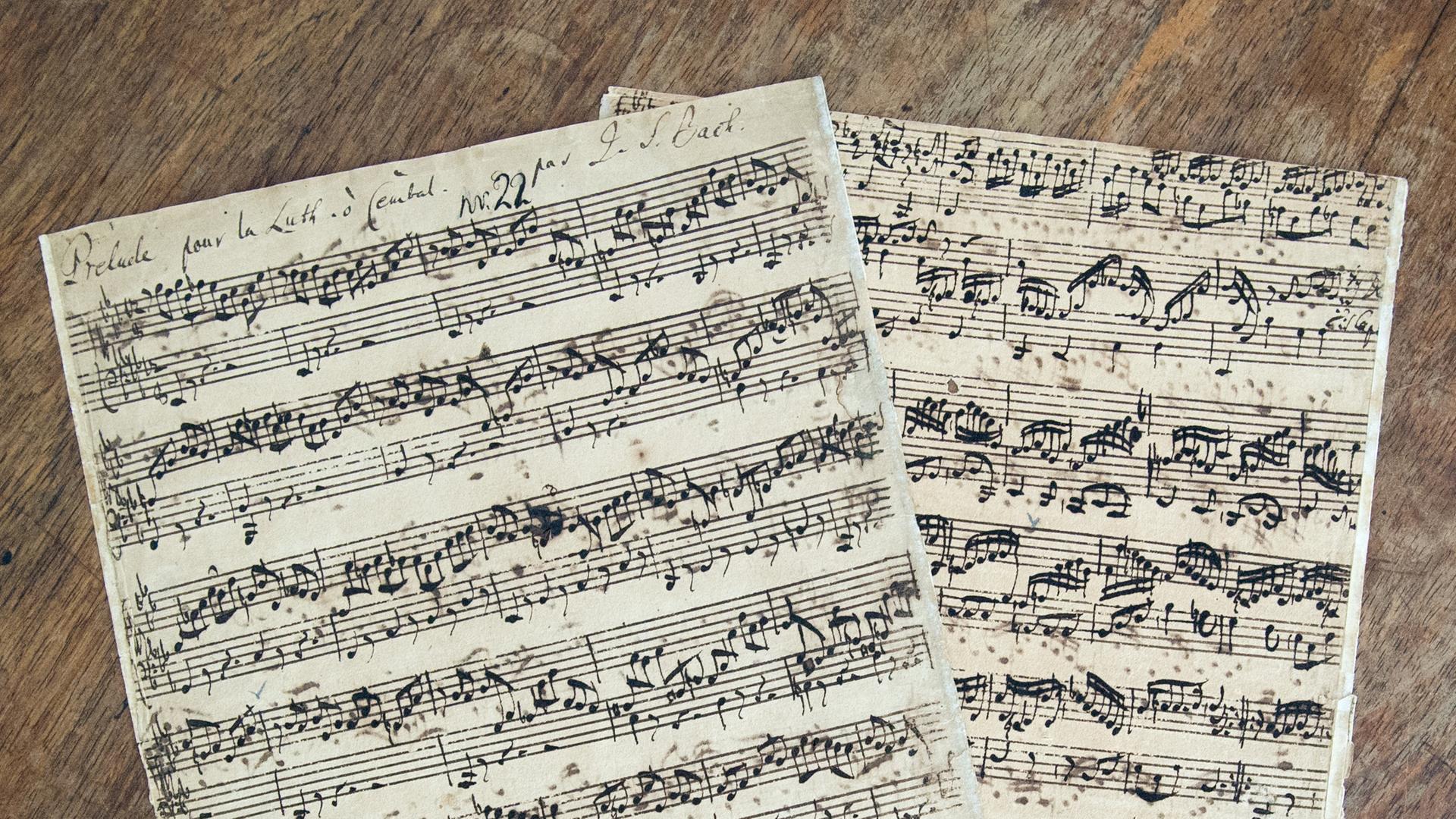 Zwei original beschriebene Notenblätter von Johann Sebastian Bach liegen übereinander gelegt auf einem Holztisch.