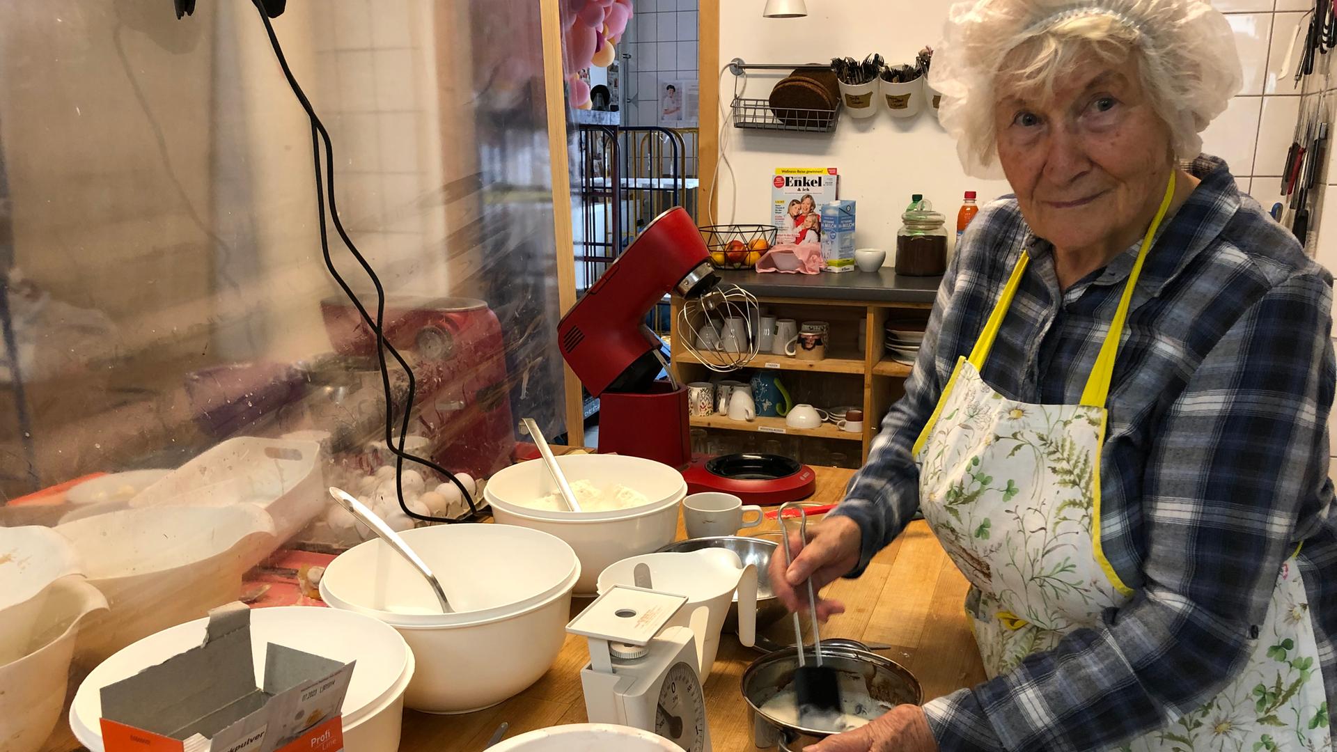 Oma Rosemarie trägt ein Haarnetz in der Küche. Vor sich hat sie eine Schüssel, in der sie etwas anrührt. Auf dem Tisch stehen weitere Schüsseln und eine Waage.