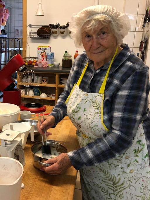 Oma Rosemarie trägt ein Haarnetz in der Küche. Vor sich hat sie eine Schüssel, in der sie etwas anrührt. Auf dem Tisch stehen weitere Schüsseln und eine Waage.