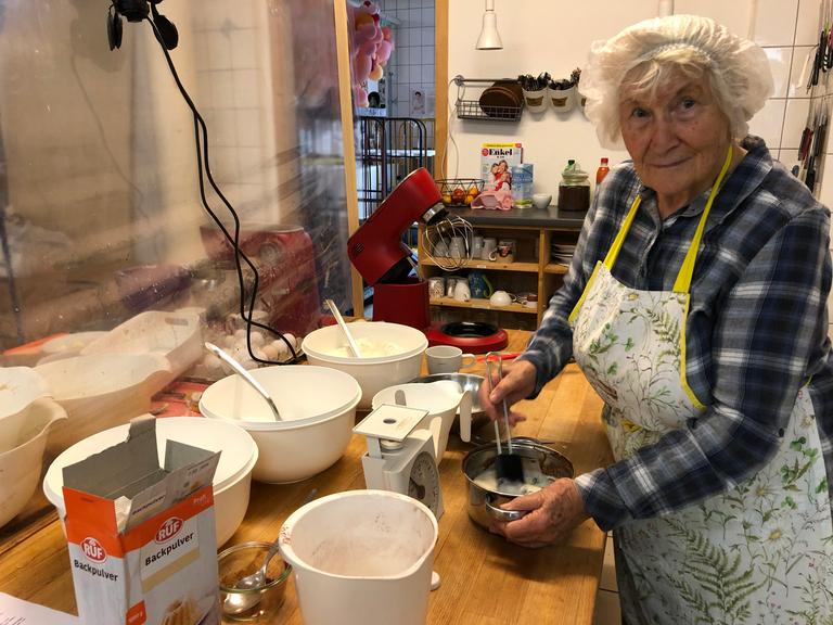 Oma Rosemarie trägt ein Haarnetz in der Küche. Vor sich hat sie eine Schüssel, in der sie etwas anrührt. Auf dem Tisch stehen weitere Schüsseln und eine Waage.