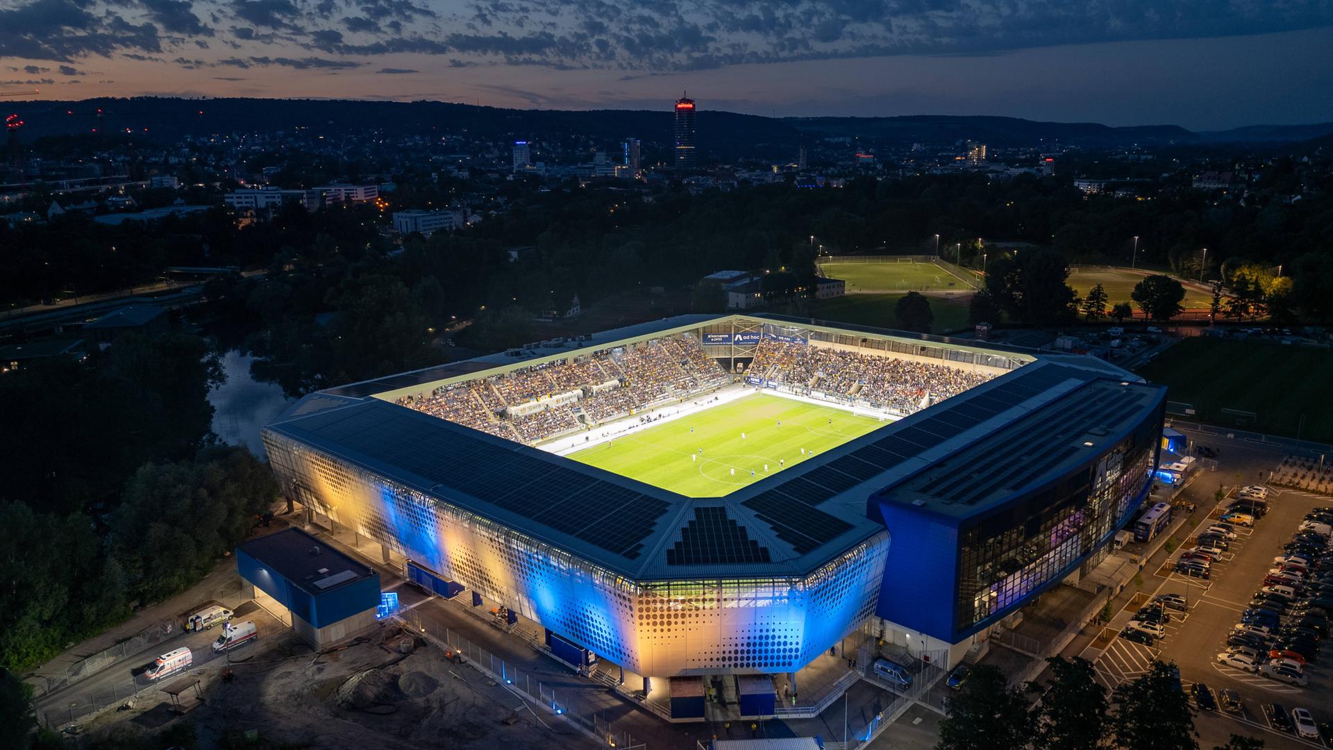 Blick aus der Luft auf das Stadion mit blau-gelb beleuchteten Seiten. Der Rasen ist hell beleuchtet. Es wird gerade Nacht. Im Hintergrund Lichter der Stadt. 