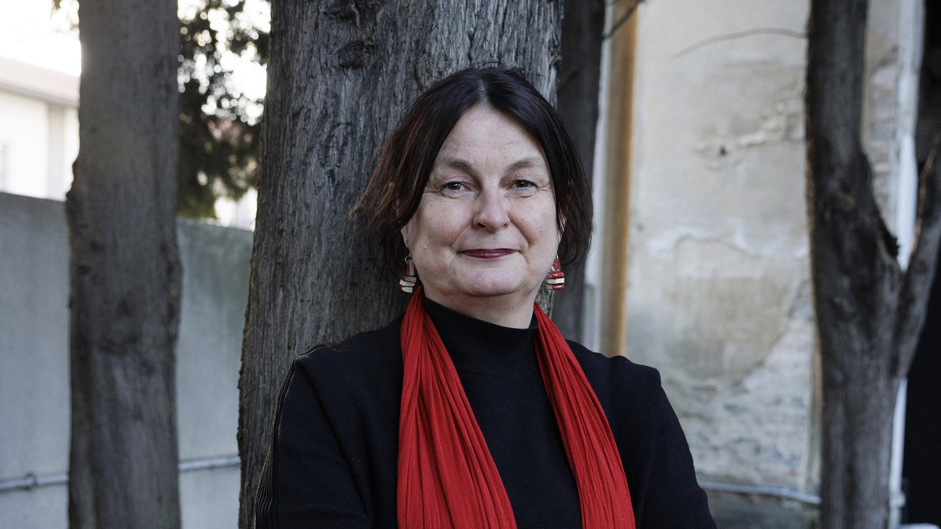 Porträtfoto einen Frau mit rotem Schal, die mit verschränkten Armen vor einem Baumstamm steht. 