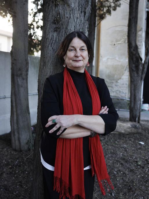 Porträtfoto einen Frau mit rotem Schal, die mit verschränkten Armen vor einem Baumstamm steht. 