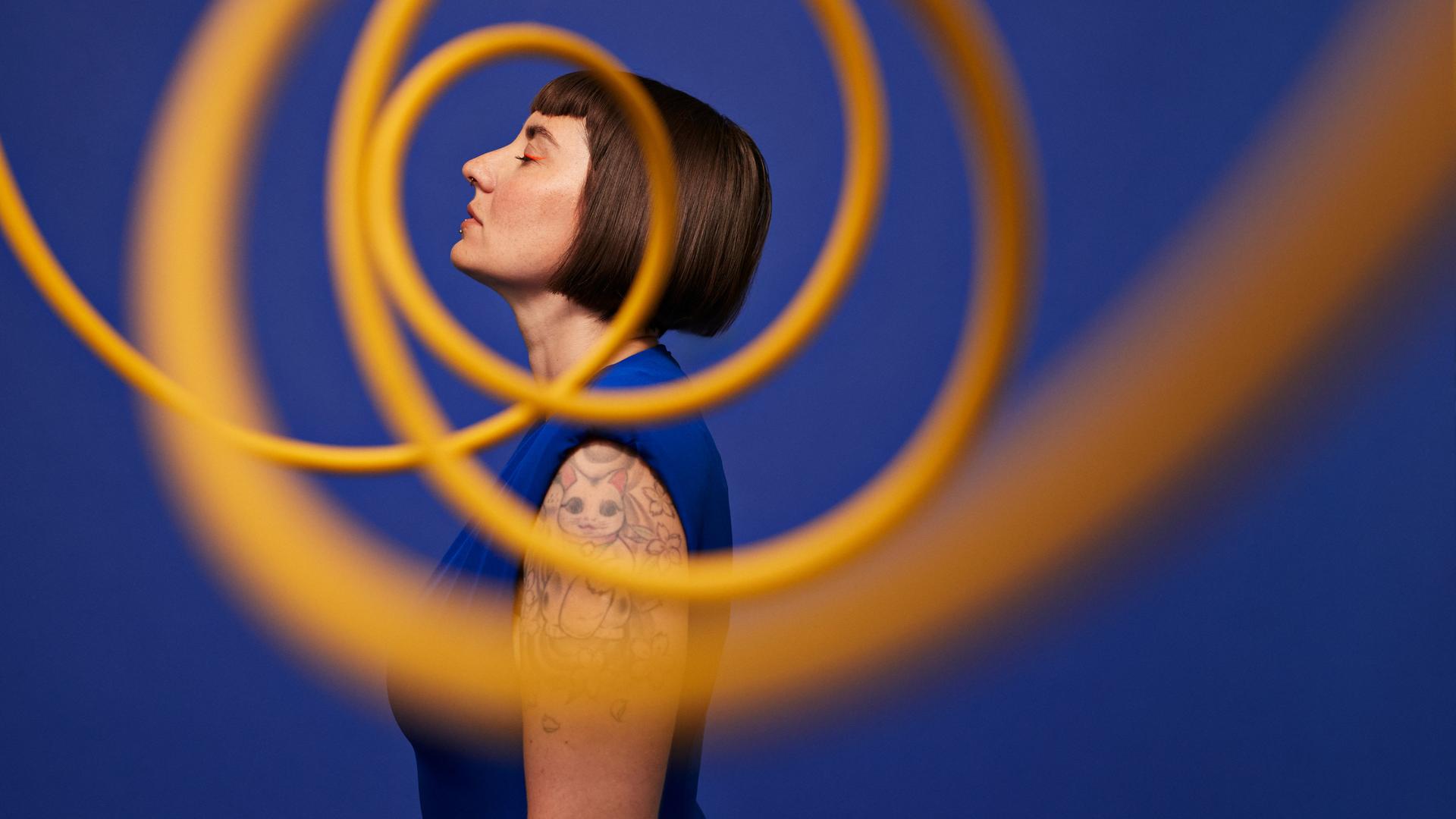 Seitenansicht einer Frau, die mit geschlossenen Augen vor blauem Hintergrund hinter gelben, in der Luft schwebenden Spiralen steht. Seitenansicht einer Frau, die mit geschlossenen Augen vor blauem Hintergrund hinter gelben, in der Luft schwebenden Spiralen steht.