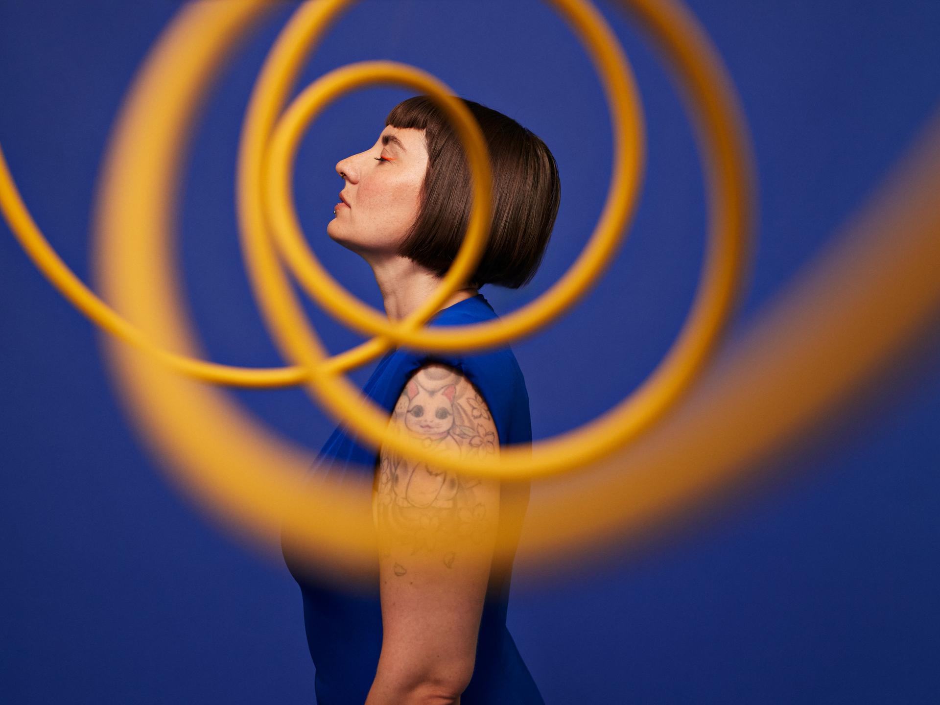 Seitenansicht einer Frau, die mit geschlossenen Augen vor blauem Hintergrund hinter gelben, in der Luft schwebenden Spiralen steht.