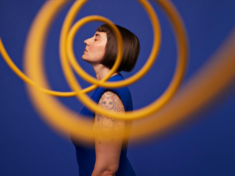 Seitenansicht einer Frau, die mit geschlossenen Augen vor blauem Hintergrund hinter gelben, in der Luft schwebenden Spiralen steht.