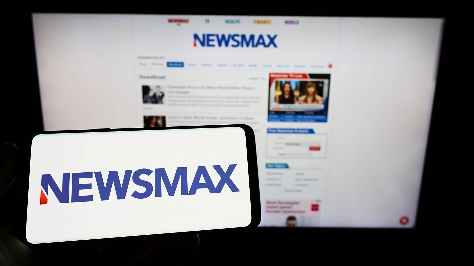Das Logo des konservativen US-Fernsehsenders Newsmax auf einem Smartphone-Bildschirm im Vordergrund sowie die Webseite von Newsmas auf einem Bildschirm im Hintergrund