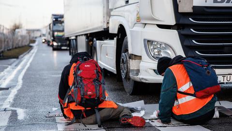 Klimaaktivisten kleben ihre Hände auf einen Zebrastreifen, so dass ein LKW von ihnen blockiert wird und sich weitere Lastwagen hinter ihm stauen.