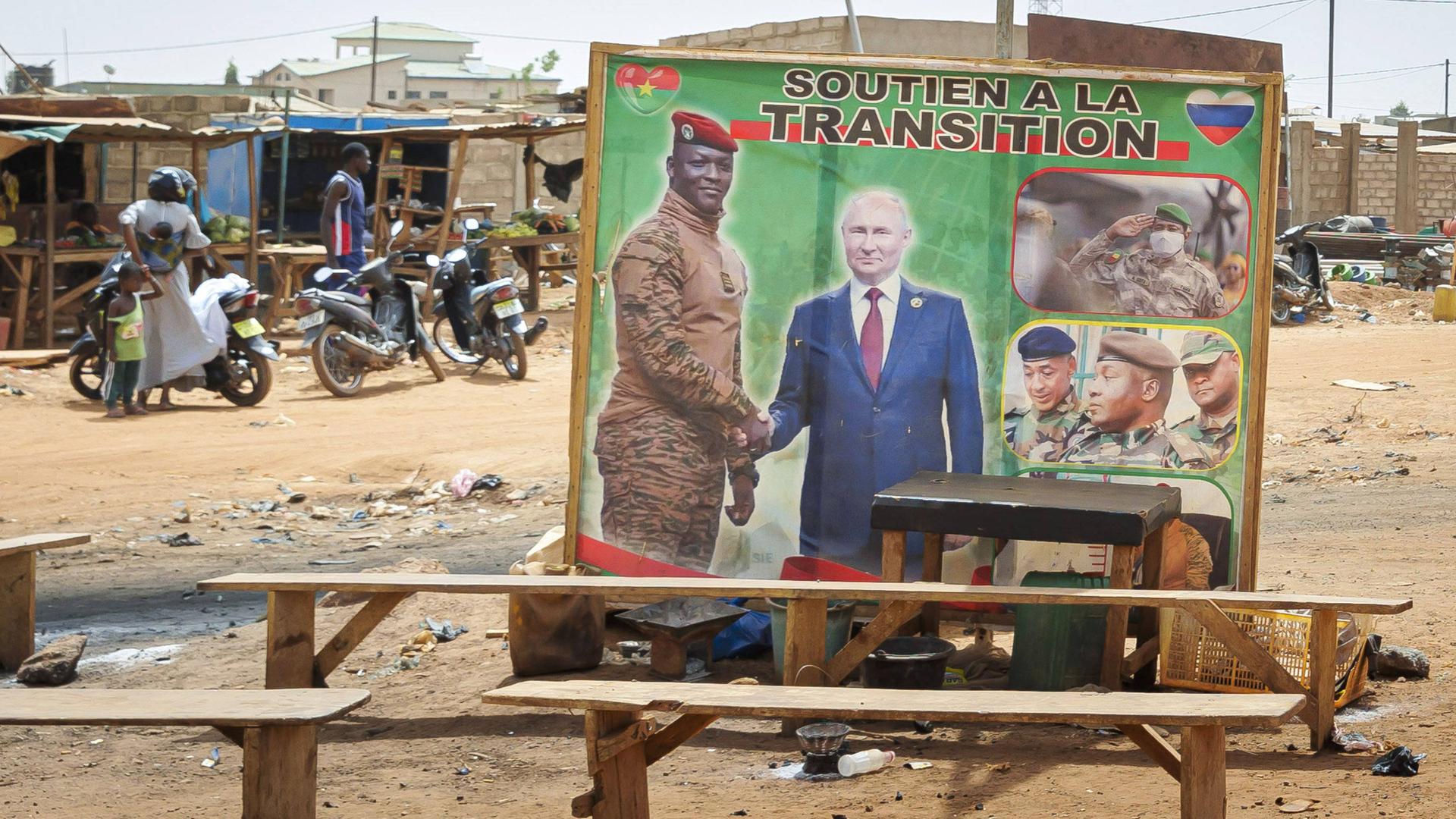 In einem Slum ohne befestigte Wege stehen mehrer Bänke herum. Dahinter ein Plakat, das zwei Männer beim Handschlag zeigt. Darüber weht eine Fahne.