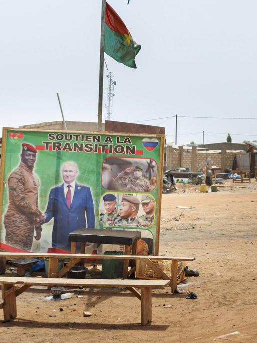 In einem Slum ohne befestigte Wege stehen mehrer Bänke herum. Dahinter ein Plakat, das zwei Männer beim Handschlag zeigt. Darüber weht eine Fahne.