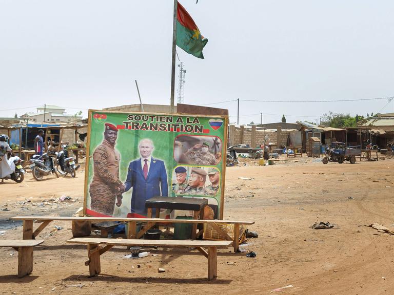 In einem Slum ohne befestigte Wege stehen mehrer Bänke herum. Dahinter ein Plakat, das zwei Männer beim Handschlag zeigt. Darüber weht eine Fahne.