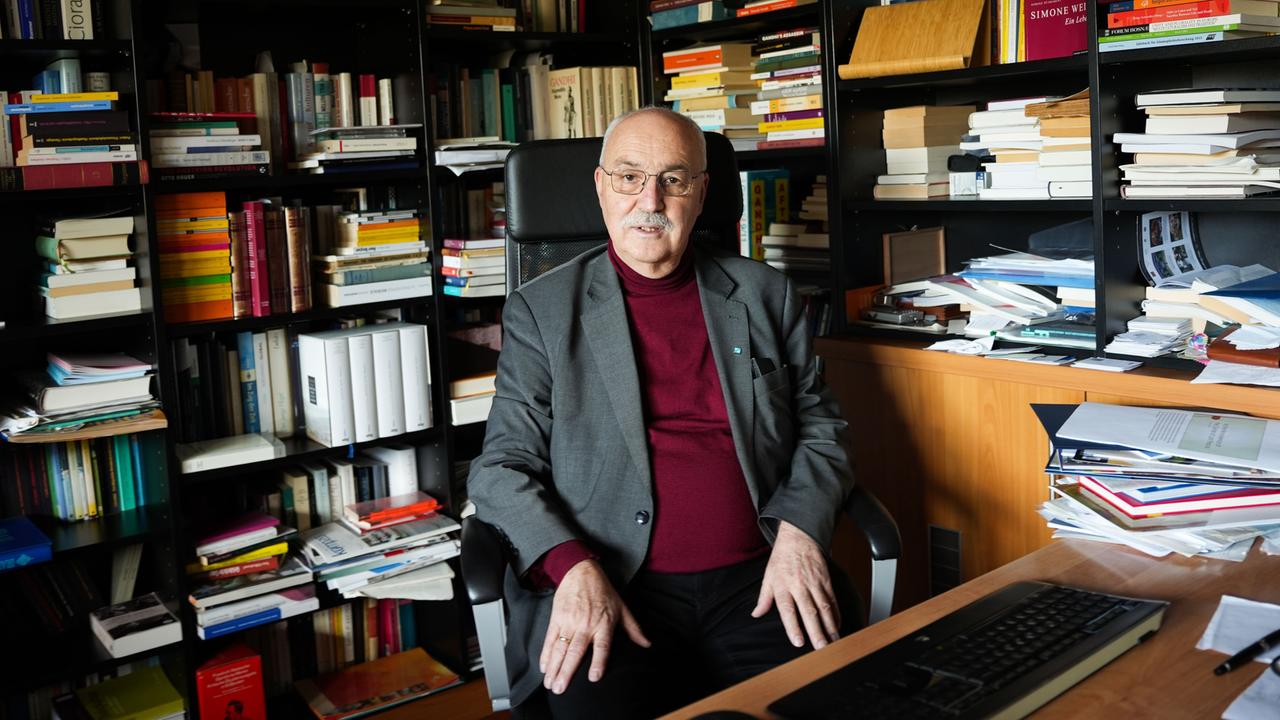 Wolfgang Palaver sitzt an einem Schreibtisch. Hinter ihm sind über Eck volle Bücherregale. 