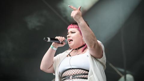 Die Rapperin Finna steht bei einem Auftritt auf der Bühne und hebt dabei den Arm mit ausgestrecktem Zeigefinger. In der anderen Hand hält sie das Mikro. Ihr Pony ist pinkfarben. Sie trägt weiße enge Kleidung und Schmuck.