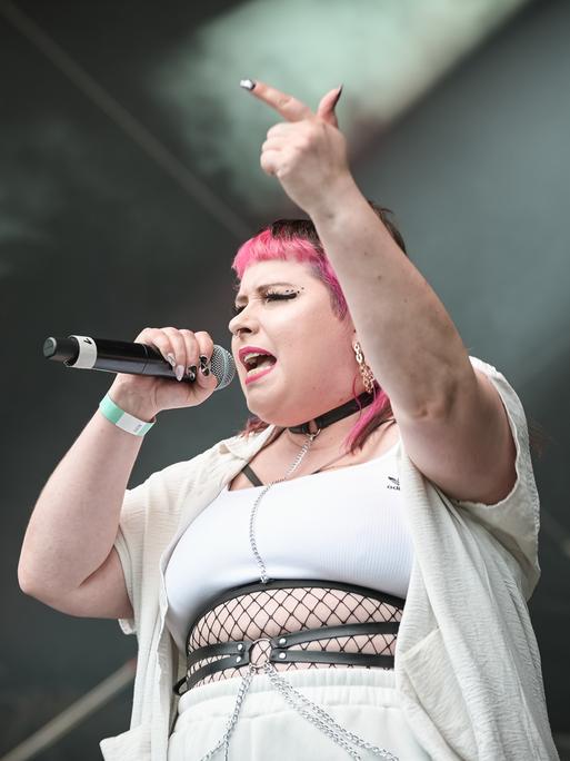 Die Rapperin Finna steht bei einem Auftritt auf der Bühne und hebt dabei den Arm mit ausgestrecktem Zeigefinger. In der anderen Hand hält sie das Mikro. Ihr Pony ist pinkfarben. Sie trägt weiße enge Kleidung und Schmuck.
