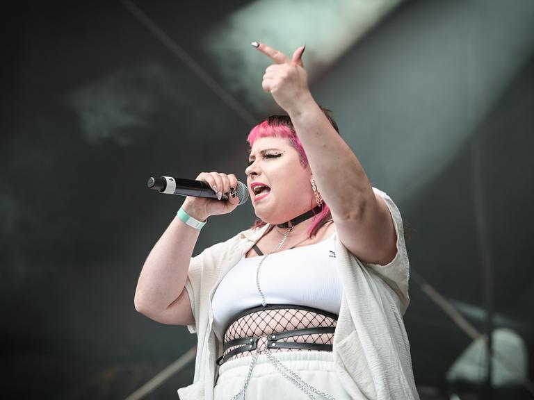Die Rapperin Finna steht bei einem Auftritt auf der Bühne und hebt dabei den Arm mit ausgestrecktem Zeigefinger. In der anderen Hand hält sie das Mikro. Ihr Pony ist pinkfarben. Sie trägt weiße enge Kleidung und Schmuck.