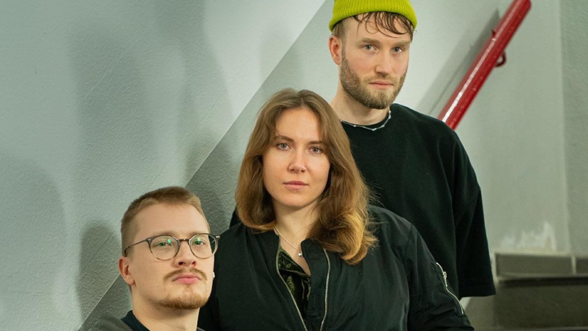 Zwei Männer stehen mit einer Frau auf einer Treppe mit rotem Geländer und schauen ernst in die Kamera.