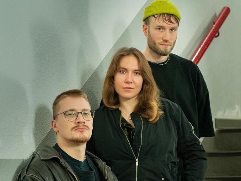 Zwei Männer stehen mit einer Frau auf einer Treppe mit rotem Geländer und schauen ernst in die Kamera.