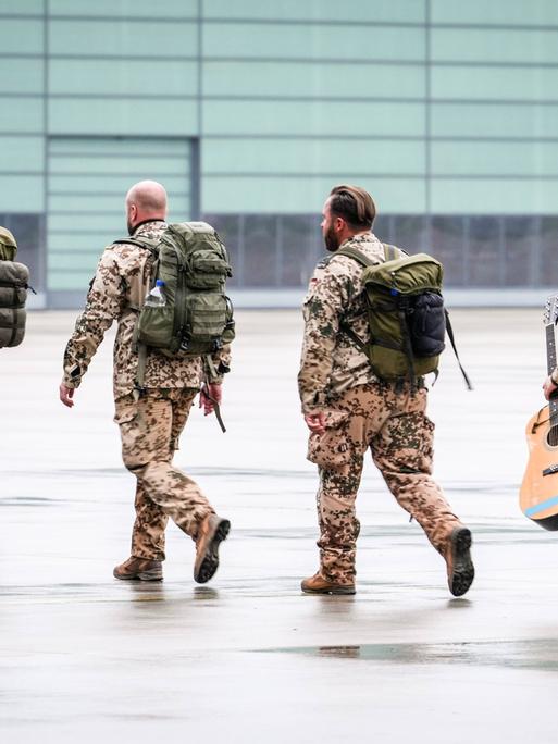 Vier Menschen in Uniformen gehen über ein Rollfeld. Sie tragen Rucksäcke. Die letzte Person ist weiblich und trägt zusätzlich eine Gitarre.