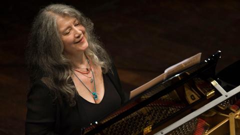 Blick von schräg oben auf die Pianistin Martha Argerich, die am Flügel sitzt mit offenem Haar und freundlich schaut, während sie in die Noten blickt und spielt. 