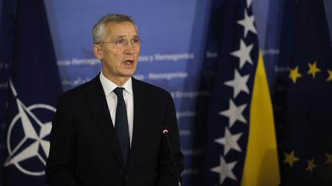 NATO-Generalsekretär Stoltenberg steht vor einem blauen Hintergrund. Die NATO und die Europa-Fahne stehen jeweils links und rechts von ihm.