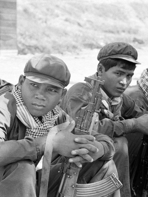 Historisches Schwarzweißfoto von Soldaten der Roten Khmer mit charakteristischen Schals, die mit ihren Waffen in einer Gruppe auf dem Boden sitzen.