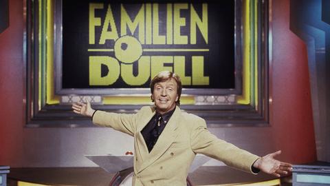 Moderator Werner Schulze-Erdel in einem beigen Sakko mit einladend ausgebreiteten Armen in der Kulisse der RTL-Quizshow "Familienduell", deren Titel hinter ihm in goldenen Lettern auf einem großen Bildschirm steht.