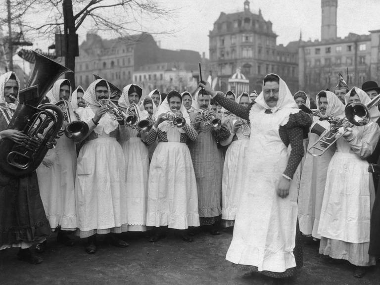 Das Orchester der "Kölner Milchmädchen" tritt in Verkleidung beim Kölner Strassenkarneval auf, ca. 1910. Eine Männerguppe in Frauenkleidung mit Kopftüchern spielt mit Blasinstrumenten einen Tusch.