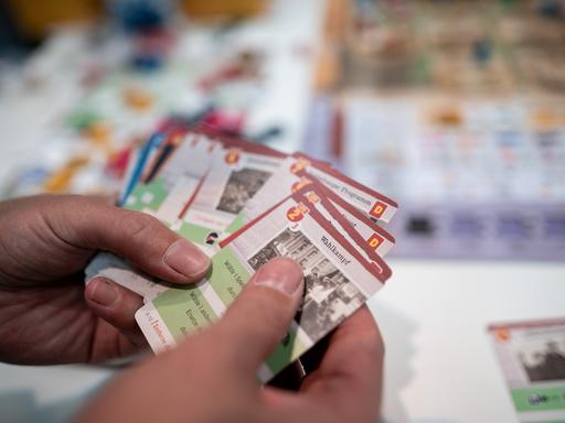 Hände halten Karten in der Hand, im Hintergrund ist ein Tisch mit einem Spielbrett und Figuren.