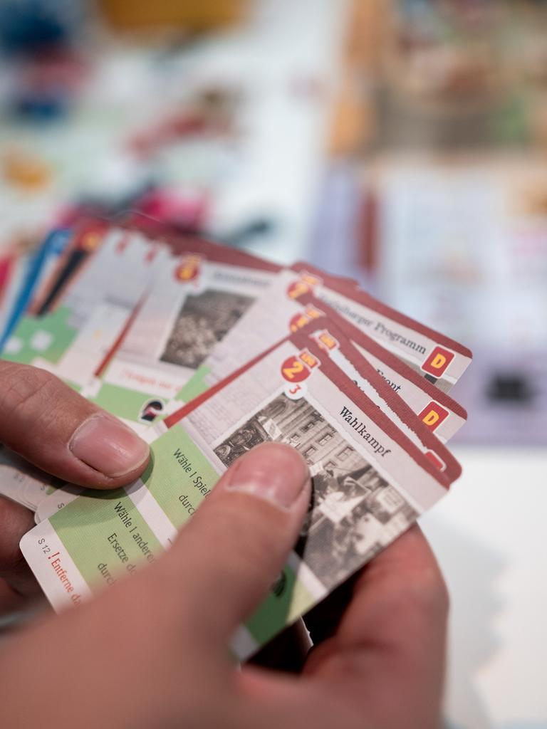 Hände halten Karten in der Hand, im Hintergrund ist ein Tisch mit einem Spielbrett und Figuren.