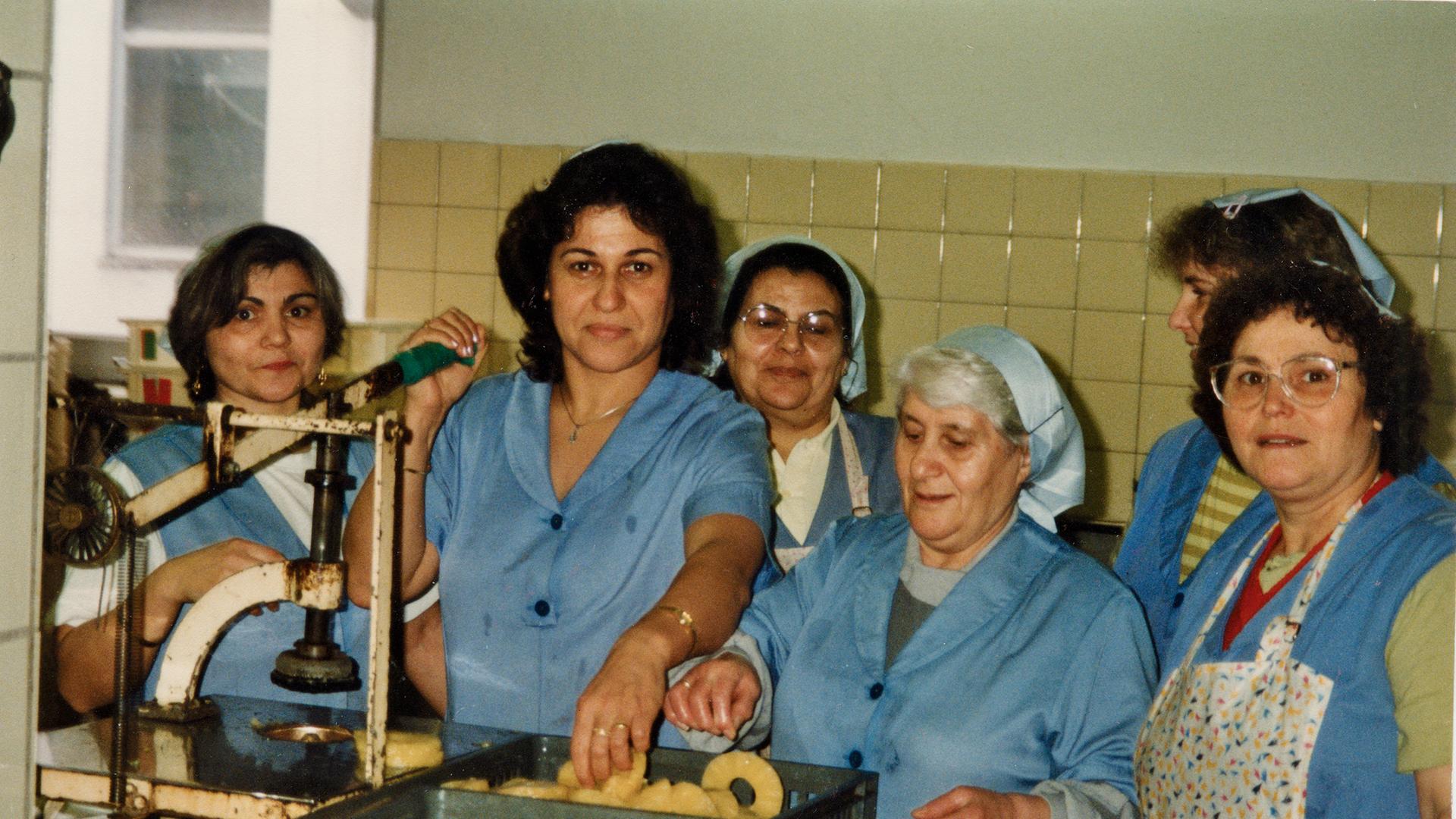 Frauen stehen in blauer Arbeitskleidung an einer Maschine. Arbeiterinnen bei Hachez.