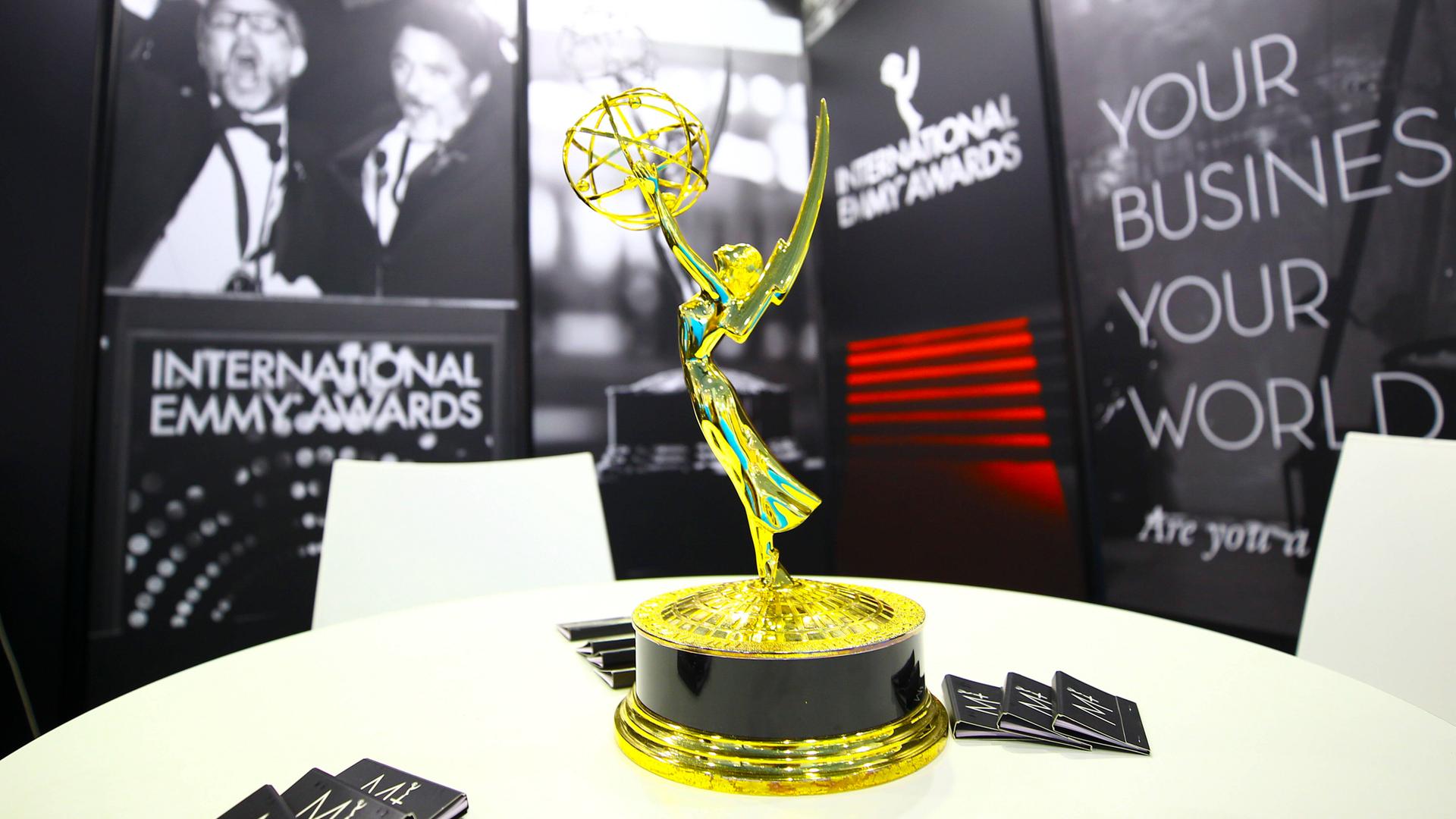 Die Trophäe des International Emmy Awards steht auf einem Tisch. Im Hintergrund sind schwarz-weiße Poster und auch der Schriftzug "Internationl Emmy Awards" zu sehen. 