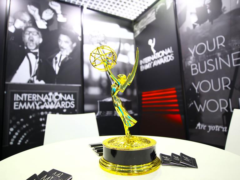 Die Trophäe des International Emmy Awards steht auf einem Tisch. Im Hintergrund sind schwarz-weiße Poster und auch der Schriftzug "Internationl Emmy Awards" zu sehen. Die Trophäe des International Emmy Awards steht auf einem Tisch. Im Hintergrund sind schwarz-weiße Poster und auch der Schriftzug "Internationl Emmy Awards" zu sehen.