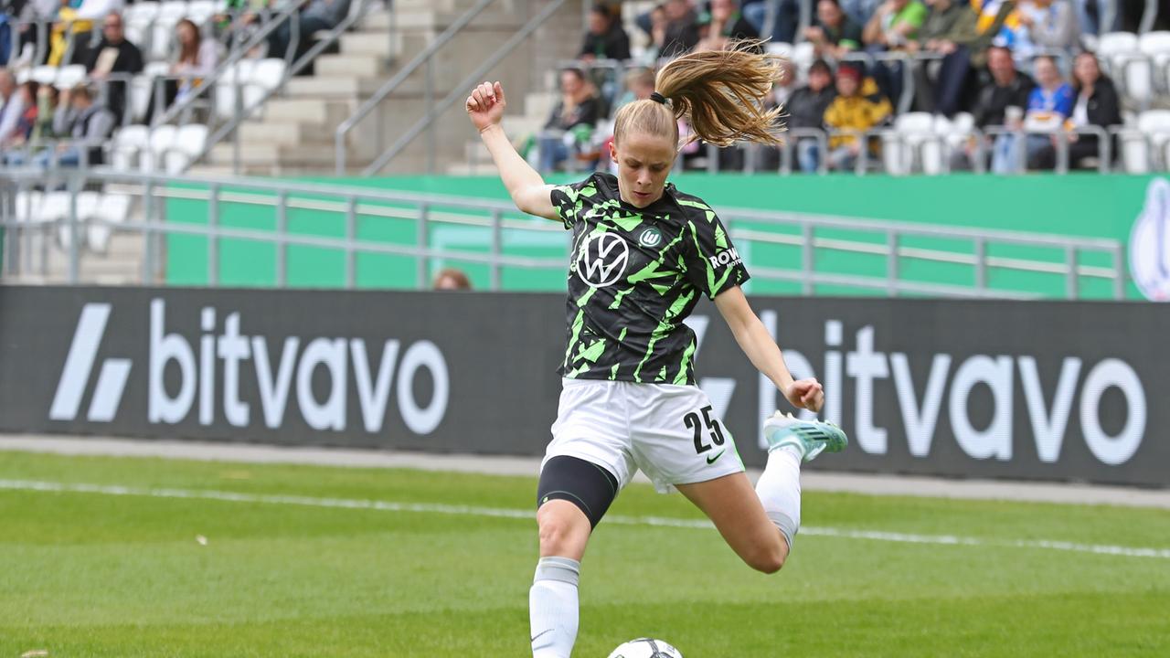 Vivien Endemann im grünen Trikot mit Applikationen holt gerade zum Schuss aus. Dahinter eine Tribüne mit Zuschauern.