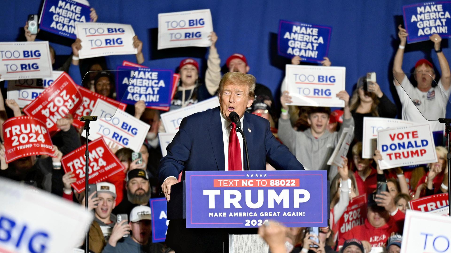 Trump steht an einem Rednerpult inmitten seiner Anhänger, die Schilder mit Aufschriften hochhalten .