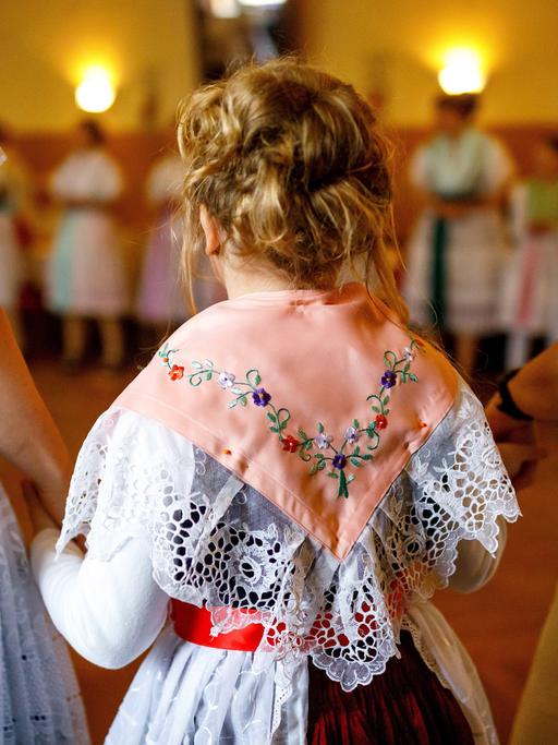 Frauen in sorbischer (wendischer) Tracht singen und tanzen im Dorfgasthof. Im Bild sind zwei Frauen von hinten zu sehen, die zwischen sich ein Kind an den Händen halten. Sie tragen traditionell bestickte Trachten.