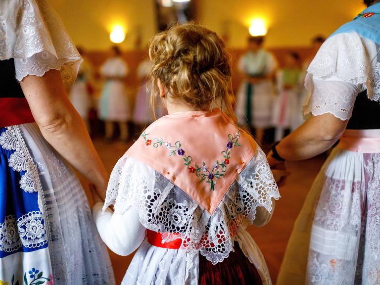 Frauen in sorbischer (wendischer) Tracht singen und tanzen im Dorfgasthof. Im Bild sind zwei Frauen von hinten zu sehen, die zwischen sich ein Kind an den Händen halten. Sie tragen traditionell bestickte Trachten.