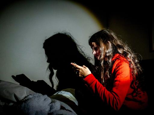Ein Mädchen im roten Pulli blickt auf ihr Handy, vor ihr der eigene Schatten. 