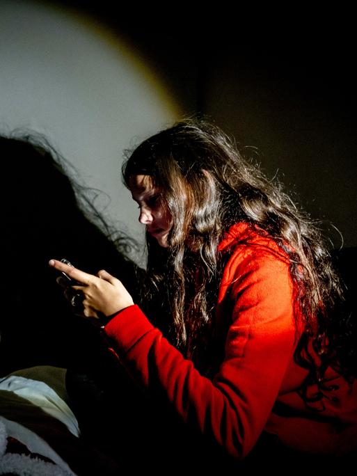 Ein Mädchen im roten Pulli blickt auf ihr Handy, vor ihr der eigene Schatten. 