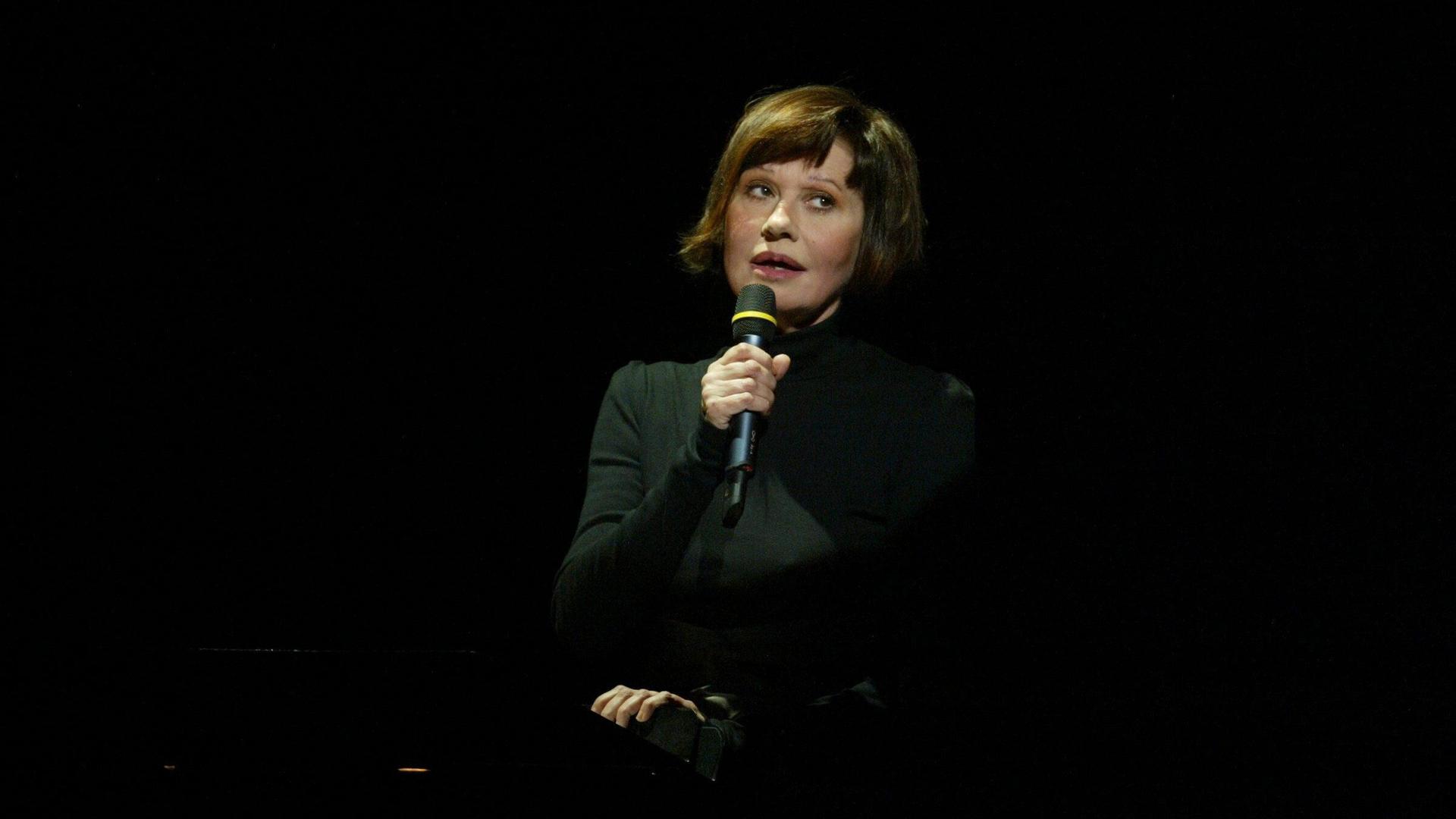 Guesch Patti sitzt auf einer dunklen Bühne und hält ein Mikrofon in der Hand. Guesch Patti sitzt auf einer dunklen Bühne und hält ein Mikrofon in der Hand.
