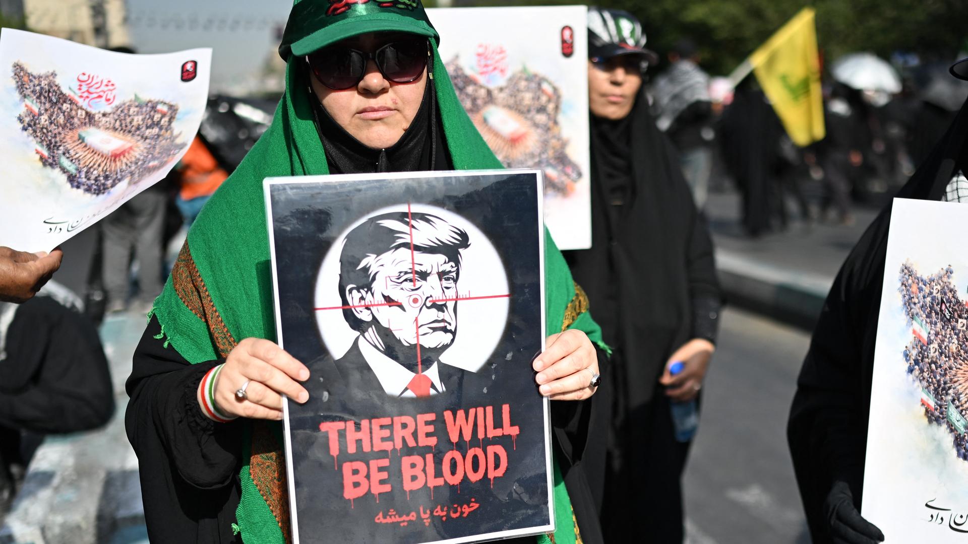 Eine Frau hält ein Plakat mit einer Illustration des US-Präsidenten Donald Trump im Fadenkreuz und der Aufschrift „There Will Be Blood“ in der Hand.