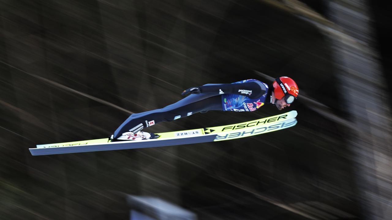 Ein Mann beim Skisprung. Er trägt einen roten Helm und einen blauen Skianzug. Er ist von der Seite fotografiert. Der Hintergrund ist verwischt.