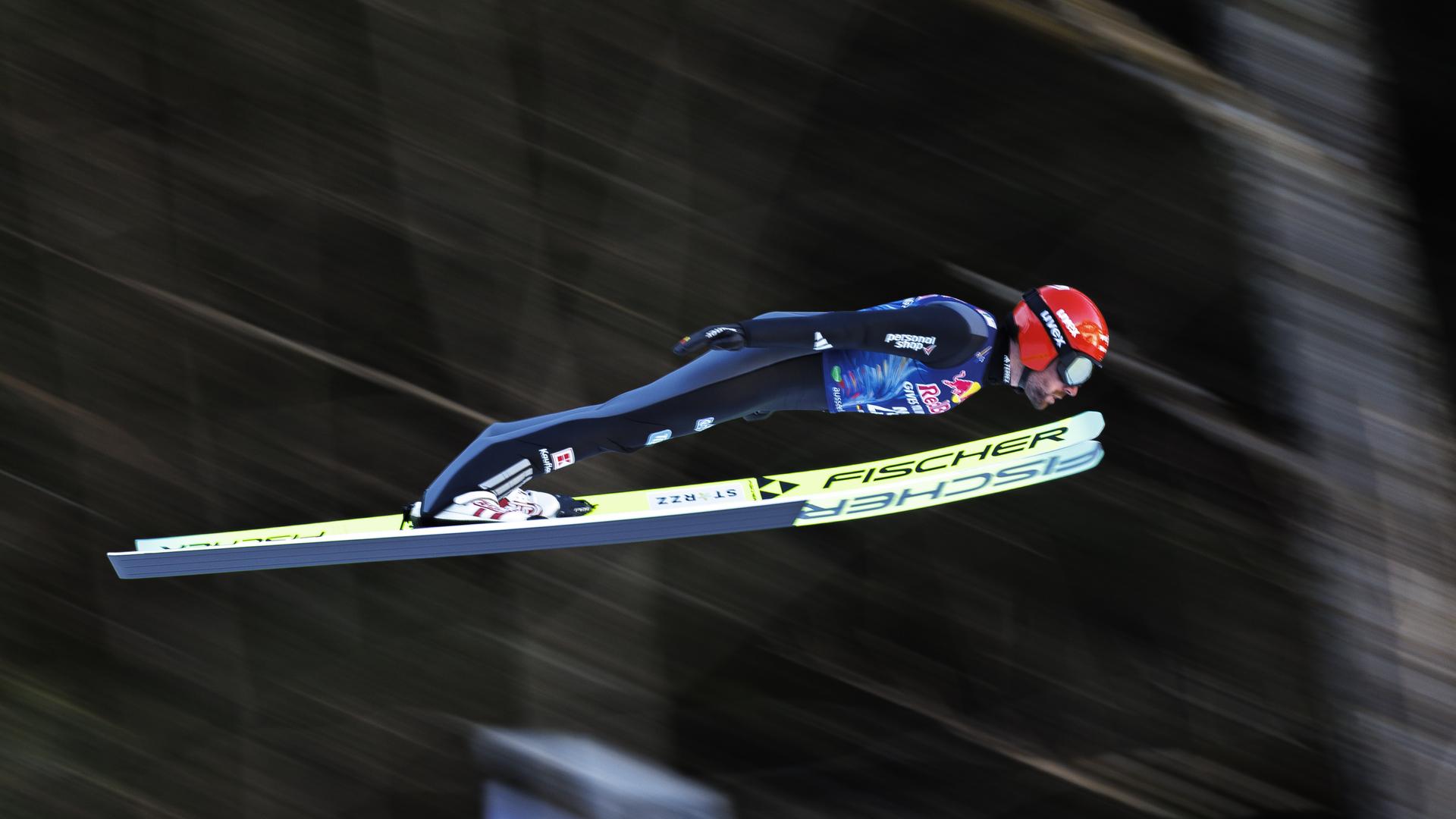 Ein Mann beim Skisprung. Er trägt einen roten Helm und einen blauen Skianzug. Er ist von der Seite fotografiert. Der Hintergrund ist verwischt.