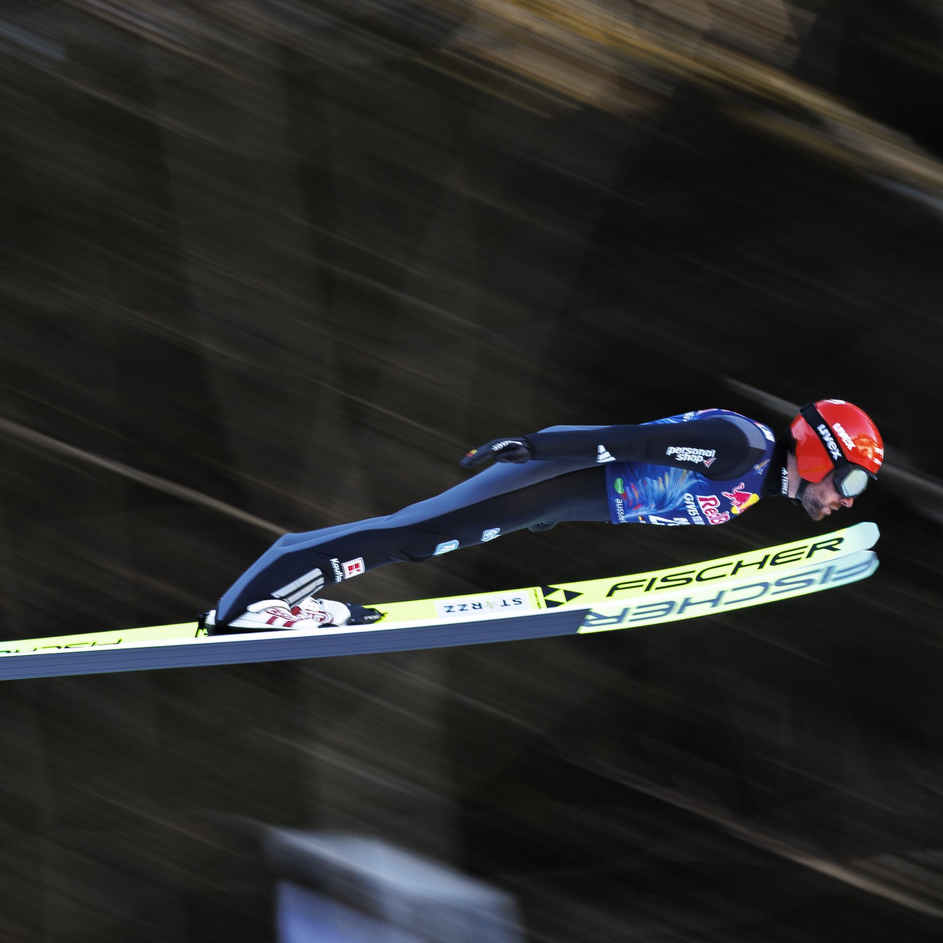 Ein Mann beim Skisprung. Er trägt einen roten Helm und einen blauen Skianzug. Er ist von der Seite fotografiert. Der Hintergrund ist verwischt.