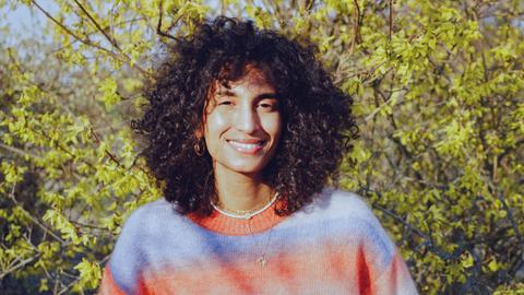 Eine junge Frau mit dunklen Locken steht vor einem gelben Forsythien-Busch. Sie lächelt in die Kamera, die Sonne scheint. Sie trägt einen blau-orange melierten Pullover.