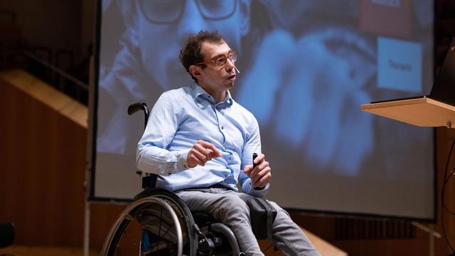 Johannes Grasser sitzt im Roll-Stuhl auf einer Bühne. Er trägt ein Mikrofon am Kopf und spricht.