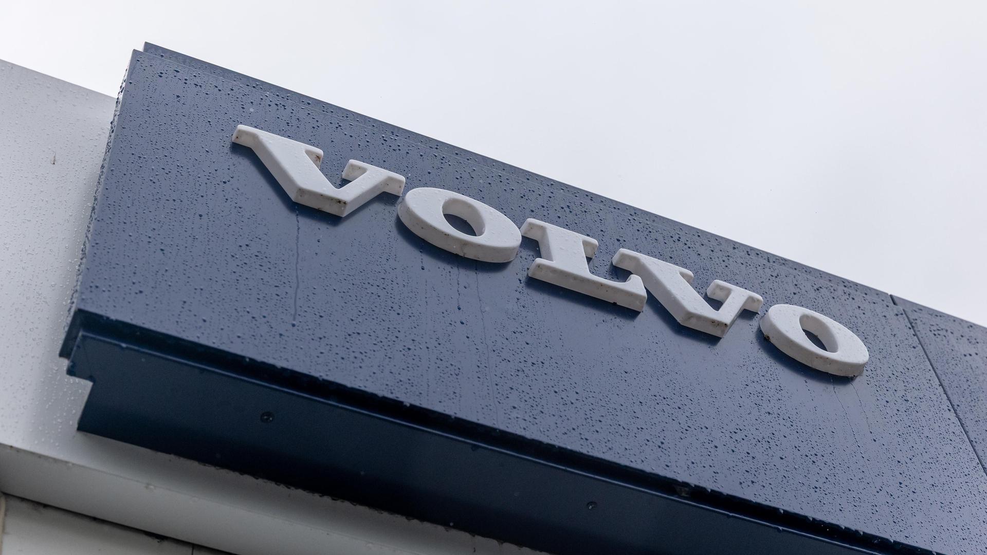 Der weiße Volvo-Schriftzug auf einem blauen Hintergrund ist an der Fassade eines Autohauses. Die Oberfläche ist von feinen Regentropfen bedeckt. 