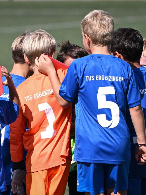 Eine Gruppe von Kindern vom TuS Erzgebirge stehen auf einem Spielfeld. Sie tragen blaue und orange Trikots und sind von hinten zu sehen. 