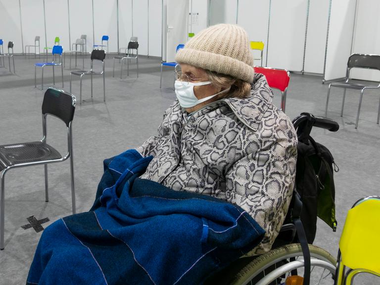 Eine ältere Fraue mit Schutzmaske und warmer Kleidung sitzt auf einem Rollstuhl in einem Impfzentrum in Spanien, 2021.